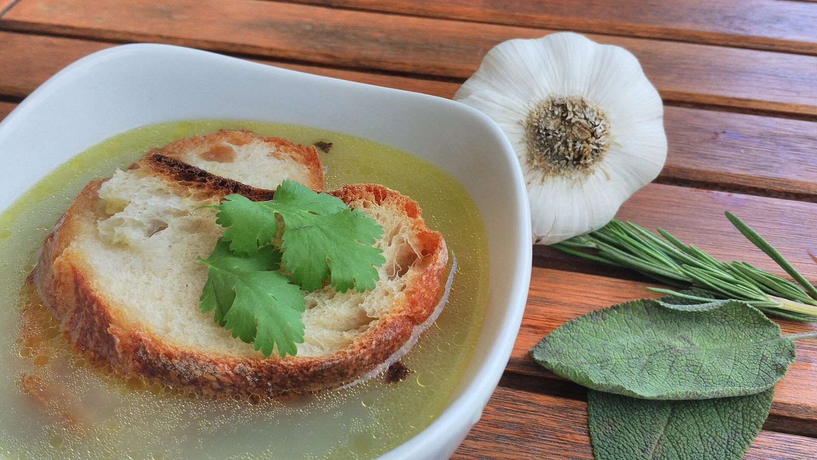 Bowl of garlic broth with a slice of toasted bread, next to a head of garlic, sage leaves and rosemary
