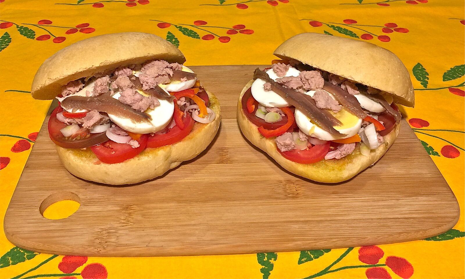 Two open pan bagnat on a wooden board, each round roll topped with tomato slices, boiled egg, flaked tuna and anchovy fillets