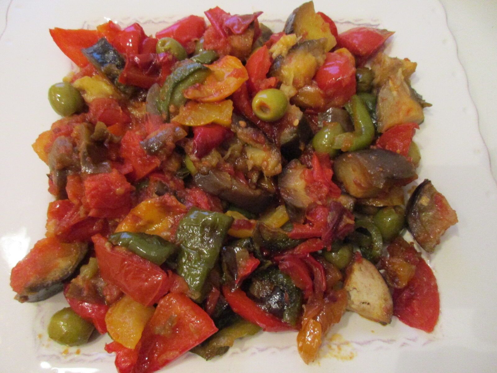 A square plate of ratatouille: chunks of tomato, red, yellow and green pepper, aubergine and whole green olives, glossy with olive oil