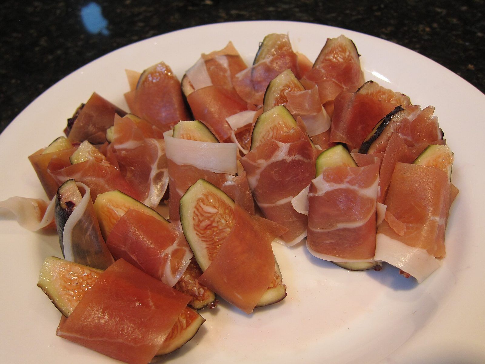 Wedges of fresh fig wrapped in thin slices of cured ham on a white plate