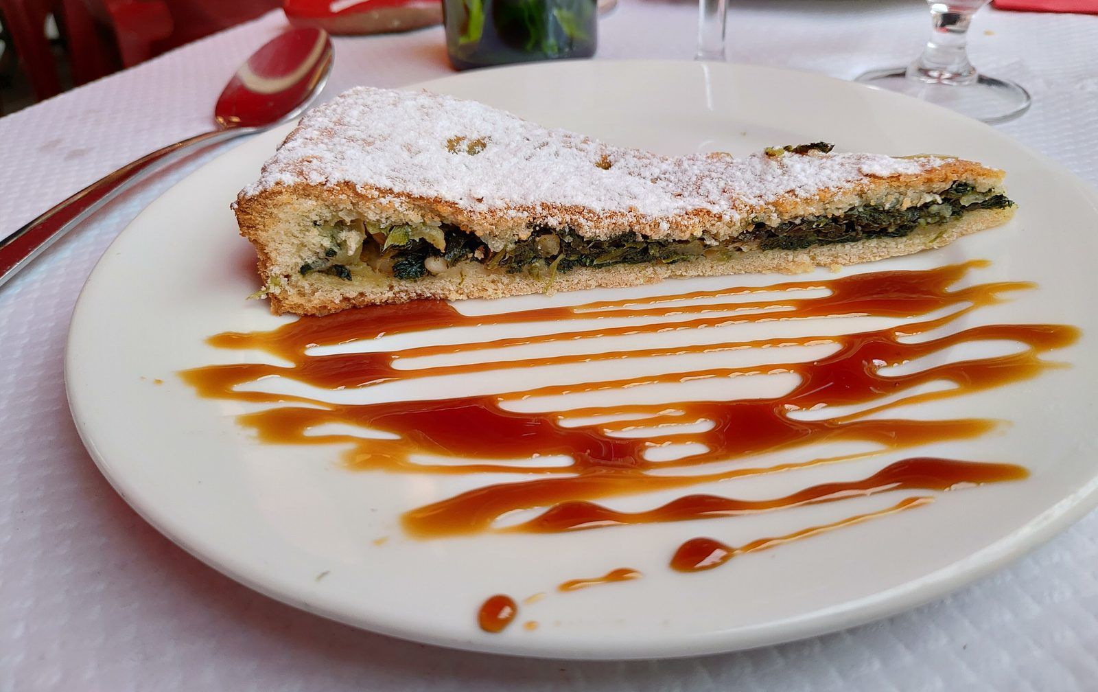 Slice of sweet Swiss chard pie with pine nuts, dusted with icing sugar, on a plate with caramel sauce